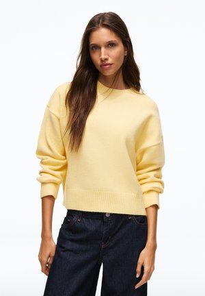 Jeune femme aux longs cheveux bruns portant un pull jaune clair et un jean bleu foncé sur un fond blanc uni.