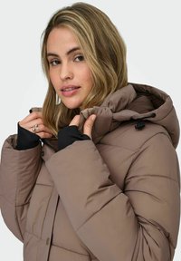 Veste rembourrée marron avec capuche, présentant un design matelassé et des accents noirs aux poignets. Le matériau semble doux et isolé.
