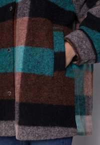 Villane ruuduline mantel, millel on türkiissinine, pruun, must ja roosa triip, nupuga kinnitamine ja küljetaskud. Pehme, tekstureeritud kangas.