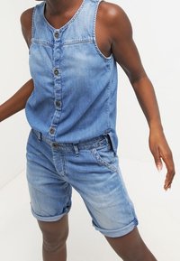 Denim overal v světlemodré barvě s bezrukávovým designem, předním zapínáním na knoflíky a ohrnutými manžetami na odpovídajících šortkách, s uvolněným střihem.