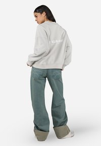Light gray oversized sweatshirt with ribbed cuffs, featuring a large white logo on the back. Paired with wide-leg green denim jeans and chunky sneakers.
