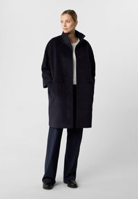 Marineblauer Wollmischmantel mit hohem Kragen, Knopfverschluss, Seitentaschen und gerippten Strickakzenten. Kombiniert mit schwarzer weitem Hosen und schwarzen Schuhen.