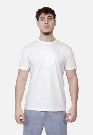 Giovane uomo con capelli corti e ricci che indossa una maglietta bianca a maniche corte semplice e jeans blu, in piedi contro uno sfondo chiaro.