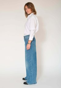 Jean en denim bleu à jambes larges et taille haute, doté d'un design classique à cinq poches et d'un léger effet usé, associé à une chemise blanche boutonnée.