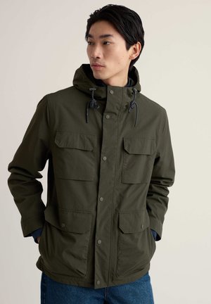 Man wearing dark green hooded jacket with four front pockets and blue jeans, standing with hands in pockets, looking left.