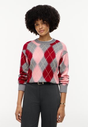Persona con capelli ricci che indossa un maglione argyle rosso, rosa e grigio e pantaloni grigio scuro, in piedi davanti a uno sfondo semplice.