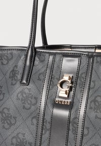 Zwarte synthetische tote bag met een reliëflogo patroon, twee stevige handvatten en een goudkleurig metalen accent met de merknaam "GUESS."