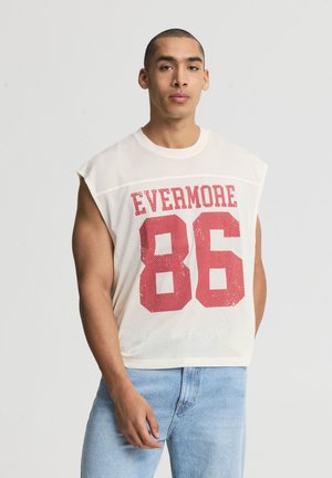 Giovane uomo con capelli corti indossa una canottiera bianca in rete con testo rosso "EVERMORE 86" e jeans azzurri, in piedi su uno sfondo semplice.