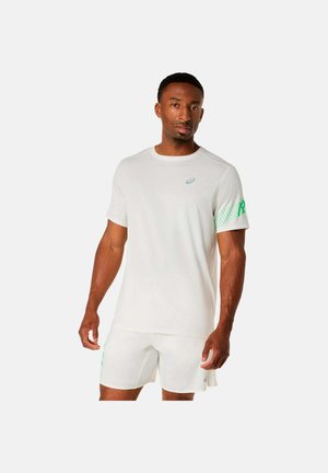 Uomo che indossa una maglietta sportiva bianca con accenti verdi sulla manica e pantaloncini bianchi abbinati, in piedi davanti a uno sfondo semplice.