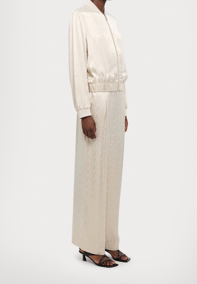 Personne portant une veste bomber en satin crème et une jupe longue assortie avec une texture subtile subtilement motifée et des sandales à talons noires, debout de côté.