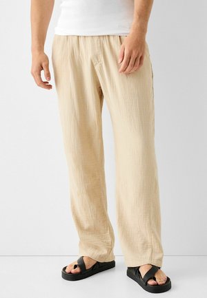 Homme portant un pantalon ample en lin beige et des sandales noires à larges brides, debout devant un fond blanc uni.
