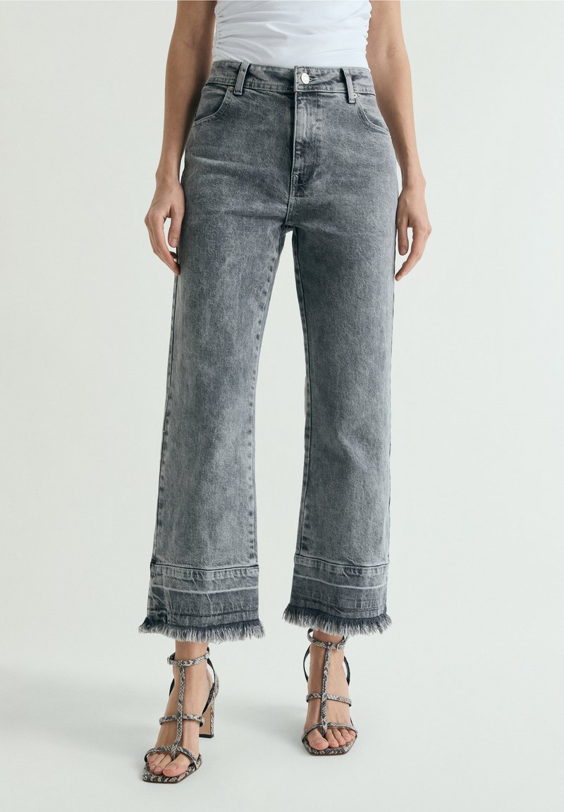 Mujer con vaqueros grises descoloridos de talle alto y tobillos deshilachados, y sandalias de tacón alto con estampado de serpiente, posando contra un fondo liso.