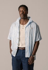 Lichtblauw shirt met korte mouwen met een golvend, wit patroon, beige gebreide onderhemd, marineblauwe broek en een bruine gevlochten riem. Accenten van goudkleurige sieraden.