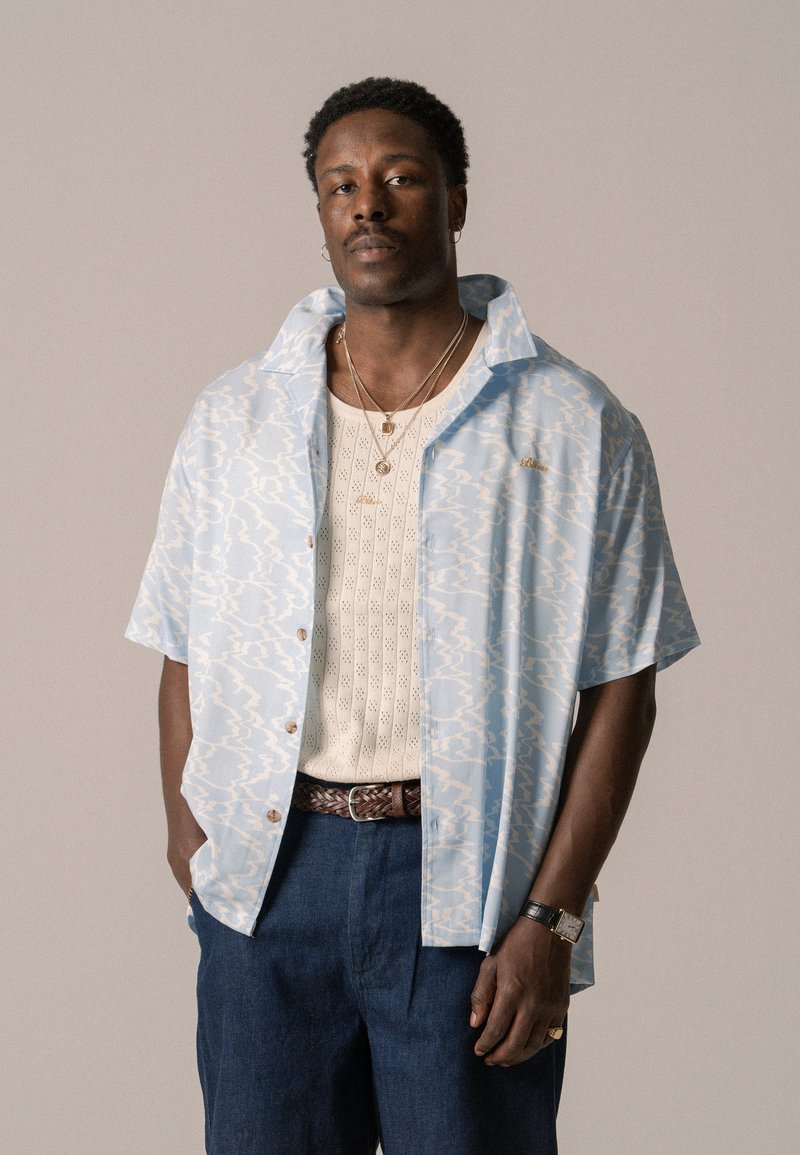 Lichtblauw shirt met korte mouwen met een golvend, wit patroon, beige gebreide onderhemd, marineblauwe broek en een bruine gevlochten riem. Accenten van goudkleurige sieraden.