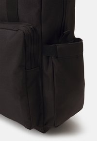 Sac à dos en tissu noir avec plusieurs poches zippées et une poche latérale. Présente une surface texturée et des bretelles réglables pour le porter.