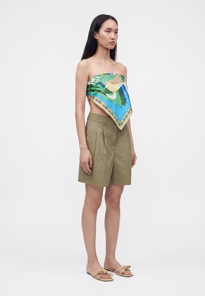 Femme debout portant un top foulard carré coloré et à motifs, un short plissé kaki, et des sandales beiges avec des détails en nœud sur un fond blanc uni.