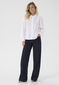 Vit blus med knappar och volangdetaljer samt långa ärmar, ihop med mörkblå vida jeans med kontrasterande sömmar.