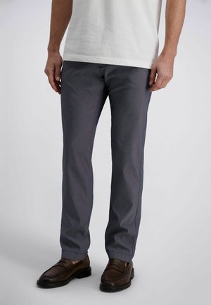 Mann trägt graue, eng anliegende Hosen, weißes Hemd und braune Leder-Loafer, steht vor einem schlichten hellen Hintergrund.