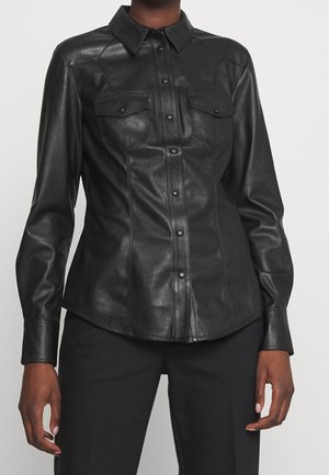 Chemise en cuir noire cintrée à manches longues avec fermeture à boutons sur le devant et deux poches à rabats sur la poitrine, portée avec un pantalon noir.