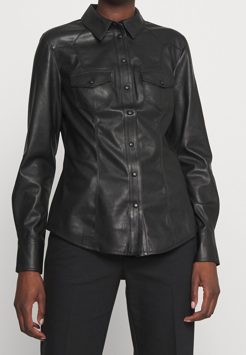 Chemise en cuir noire cintrée à manches longues avec fermeture à boutons sur le devant et deux poches à rabats sur la poitrine, portée avec un pantalon noir.