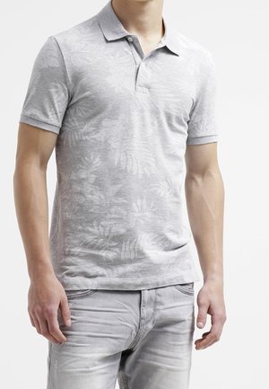 Mężczyzna ubrany w jasnoszarą koszulkę polo z krótkim rękawem w kwiatowy wzór oraz jasnoszare dżinsy, stojący z ramionami luźno opuszczonymi wzdłuż ciała.