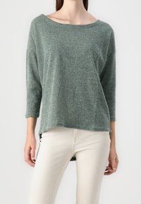 Haut en maille verte texturée avec un large col bateau et des manches trois-quarts. L'ourlet est plus long à l'arrière, associé à un jean de couleur claire.