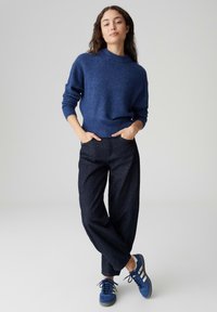 Maglione di lana blu con collo alto, abbinato a pantaloni ampi in denim scuro e sneakers blu in suede con strisce bianche.