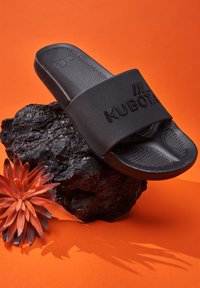 Czarna klapka nałożona z teksturowanej gumy z szerokim paskiem z wyembosowanym logo "KUBOTA", umieszczona na ciemnej, skalistej powierzchni.