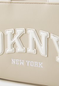 Bézs szintetikus kézitáska kiemelkedő fehér "OKNY" felirattal és hímezett "NEW YORK" szöveggel. Simaságával és kontrasztos varrásával kiemelkedő stílust képvisel.