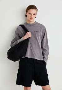 Graues langärmliges Shirt mit Rundhalsausschnitt, kombiniert mit schwarzen Shorts, hält eine schwarze, gesteppte Umhängetasche. Einfache Gestaltung, schmucklos.