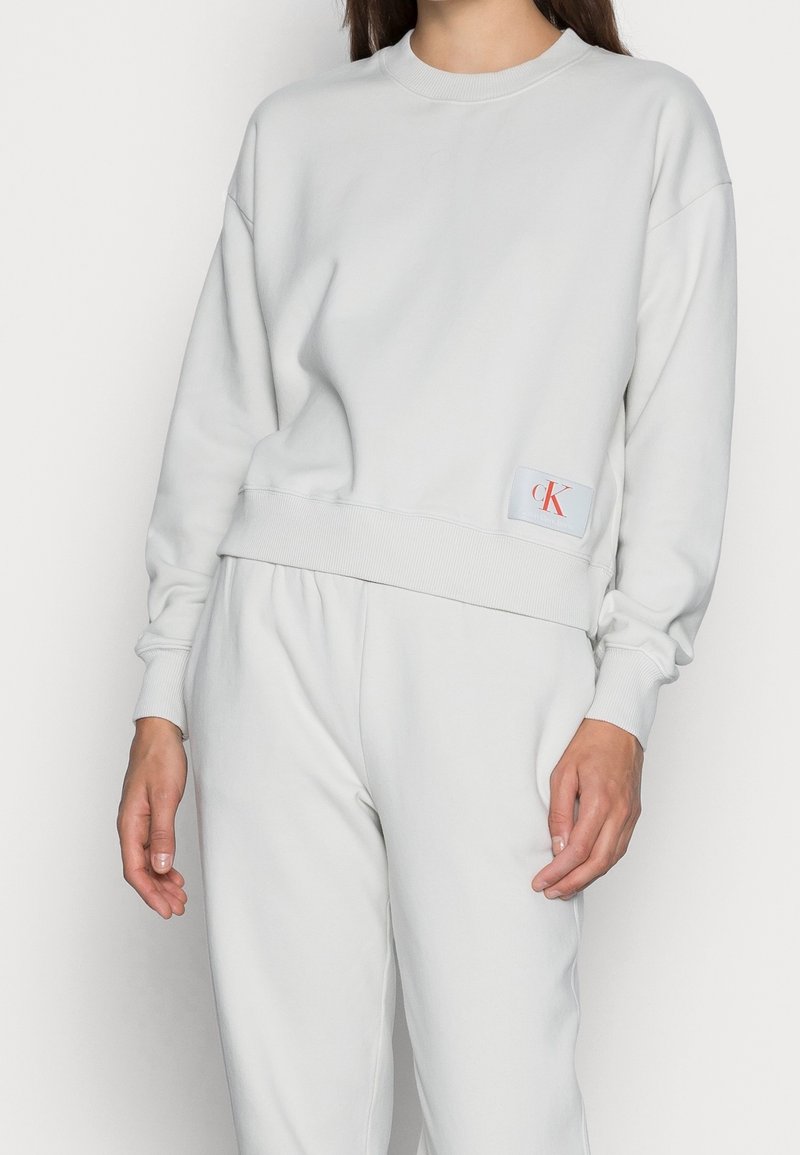 Femme portant un sweat-shirt gris clair Calvin Klein et un pantalon de survêtement assorti, debout devant un fond blanc uni.