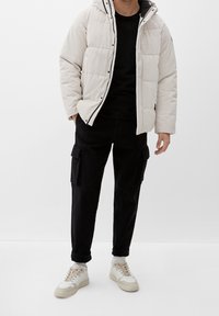 Homme portant une doudoune beige clair, un pantalon cargo noir, une chemise noire, des chaussettes blanches et des baskets blanches, debout devant un fond uni.