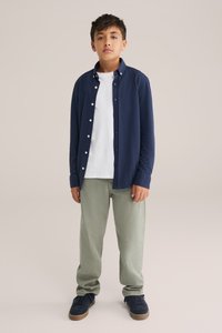 Jūras zila garām piedurknēm pogājama krekla slānis virs baltā t-kreklu, apvienots ar gaiši zaļām džinsām un jūras zilām sporta apaviem. Gluda auduma, ikdienišķs piegriezums.