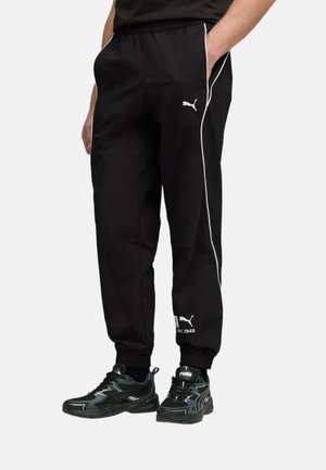 Persona che indossa pantaloni della tuta Puma neri con piping bianco, logo sulla coscia e sulla caviglia, abbinati a sneakers Puma nere.
