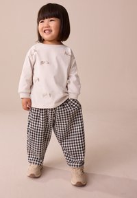 Chemise blanche à manches longues avec des accents de nœud et un pantalon à carreaux noir et blanc, associé à des baskets beige clair, sur un fond neutre.