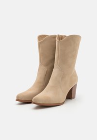Bottes basses en daim beige avec un bout pointu, un talon bloc de hauteur moyenne et un bord supérieur incurvé décoratif. Texture lisse, détails de couture minimalistes.