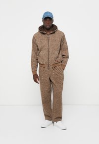 Bruin gemarmerd trainingspak met een rits hoodie en joggingsbroek, voorzien van een zachte voering, gecombineerd met een lichtblauwe pet en witte sneakers.