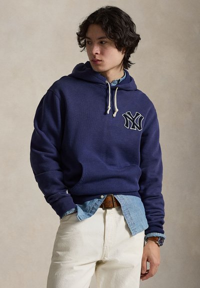 Marineblå hettegenser med hvit logo, knytinger og kengurulomme, lagd over en lyseblå skjorte og kombinert med kremfargede bukser.