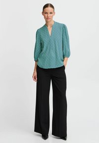 Blusa teal con scollatura a V e maniche a sbuffo, caratterizzata da un motivo delicato. Abbinata a pantaloni neri a gamba larga e scarpe nere a punta.