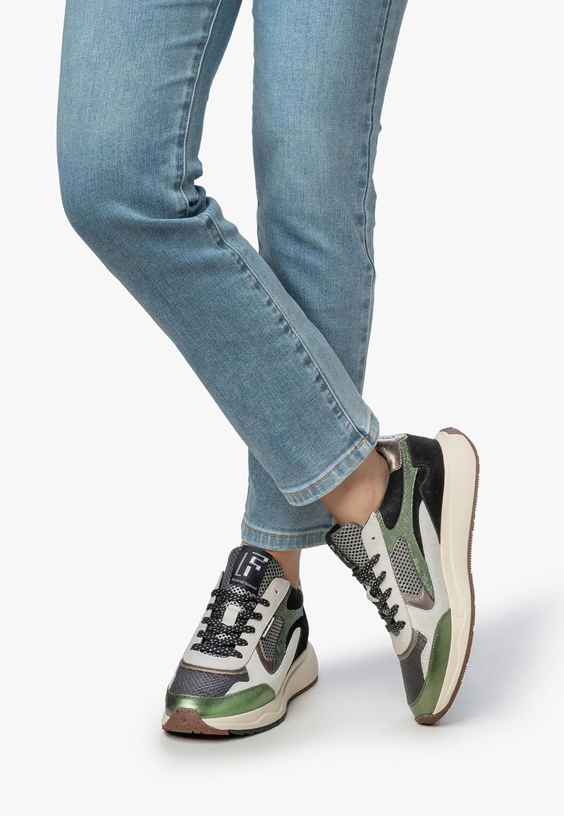 Jalat dalam jeans biru muda bersilang, mengenakan sneakers jala hijau, hitam, putih, dan abu-abu dengan tali hitam di atas latar belakang putih.
