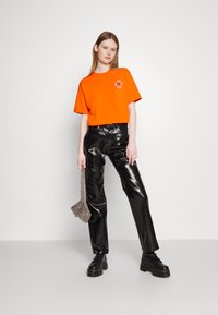 Oranje katoenen T-shirt met ronde hals en grafische print op de borst, gecombineerd met zwarte glanzende faux leren broeken en zwarte veterschoenen.