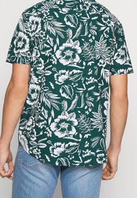 Miestenská košeľa s krátkym rukávom v tmavozelenej látke s bielymi kvetinovými a listovými vzormi, s okrúhlym spodným lemom a jednoduchým dizajnom goliera.