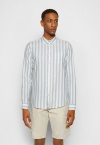 Camicia a maniche lunghe a righe blu chiaro e bianco con colletto a bottoni e taschino. Abbinata a pantaloni corti beige, in piedi contro uno sfondo neutro.
