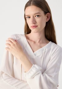 Witte, lichte blouse met een V-hals, trekkoorddetails en elastische manchetten. Vlotte textuur met een relaxte pasvorm.