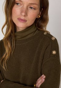 Maglione verde oliva con design a collo alto, caratterizzato da accenti in oro sui gomiti e un tessuto morbido e textures.