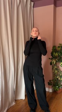 Musta kõrge kaelusega sviitri ja pükstega inimene seisab silmad kinni, kohandades kraed, kreemika kardina ja rohelise toataime lähedal pehmes roosas toas.