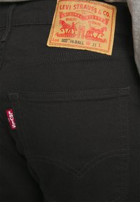 Svarta denimjeans med ett synligt orange läderpatch märkt "LEVI STRAUSS & CO." Röd "Levi's" etikett på bakfickan. Klassisk femficksdesign.