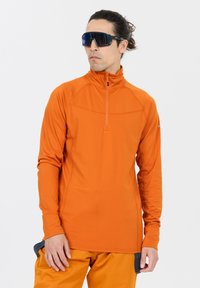 Oranger Langarm-Pullover mit einem Viertelreißverschluss, glatter Textur und taillierter Passform; kombiniert mit orangen Hosen und reflektierenden Sonnenbrillen.