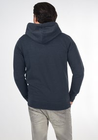 Felpa blu navy con tasca a canguro frontale, maniche lunghe e un ampio cappuccio; abbinata a jeans grigi chiari, che mostrano un look casual moderno.