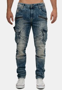 Uomo che indossa pantaloni cargo in denim blu sbiadito con tasche con zip e patta, abbinati a sneakers bianche, in piedi contro uno sfondo semplice.